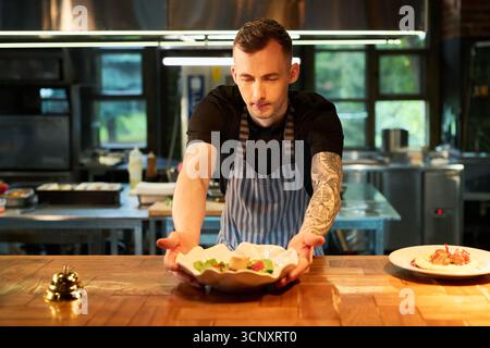 Kaukasischer junger erwachsener Mann mit tätowiertem Arm, der Gourmet-Gerichte in moderner Restaurantküche serviert, hinter der Theke steht und dem Kunden Speisen präsentiert, fokussierter Ausdruck Stockfoto