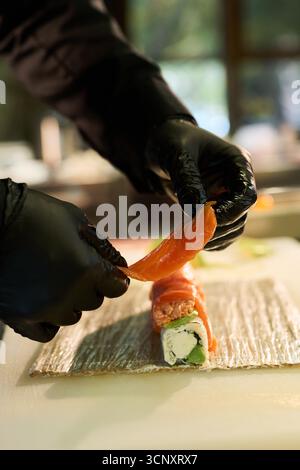 Kaukasische junge Erwachsene Hände tragen schwarze Handschuhe, bereiten Sushi-Rollen vor, legen Sie vorsichtig eine Scheibe rohen Lachses auf Reis und füllen Sie sie mit Bambus-Sushi-Matte Stockfoto