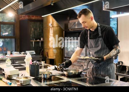 Kaukasischer junger erwachsener Mann mit tätowiertem Arm, der Lebensmittel in der professionellen Küche zubereitete, Schürze und Handschuhe trug, konzentriert sich auf den Kochprozess mit verschiedenen Utensilien und Zutaten sichtbar Stockfoto