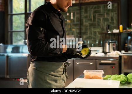 Junger Erwachsener kaukasischer Mann, der Avocado in der professionellen Küche zubereitete, in Küchenuniform und schwarzen Handschuhen, an der Theke mit Lebensmittelzutaten stand und sich auf kulinarische Aufgaben konzentrierte Stockfoto