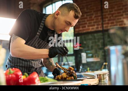 Junger Erwachsener kaukasischer Mann Koch, der Gourmet-Gericht in der professionellen Küche zubereitet, mit Pinzette Essen auf Teller anordnen, fokussierter Ausdruck, Backsteinwand Hintergrund sichtbar Stockfoto