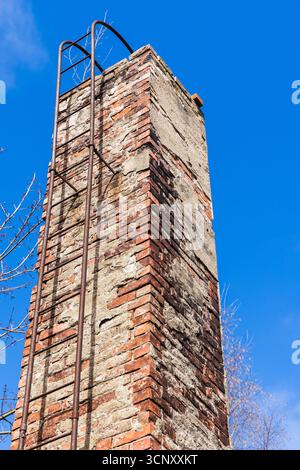 Ein verwitterter Ziegelkamin erhebt sich gegen einen leuchtend blauen Himmel, gestützt von einer verrosteten Metallleiter. Die Szene erinnert an Verlassenheit, Widerstandsfähigkeit und rustikalen Bogen Stockfoto