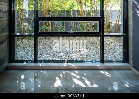 Ein elegantes, minimalistisches Zimmer mit großem Fenster und Blick auf einen Schotterhof mit Bambuspflanzen, eingerahmt von den Schatten der Blätter. Stockfoto