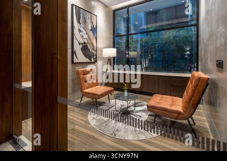 Ein gemütlicher und stilvoller, moderner Loungebereich mit zwei Ledersesseln, einem kleinen Marmortisch und einem abstrakten Gemälde, alle in das warme Licht eines Bodens getaucht. Stockfoto