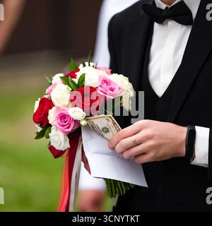 Nahaufnahme eines Bräutigams in einem Smoking mit einem wunderschönen Blumenstrauß aus roten, rosa und weißen Rosen. Ein kleiner Geldbetrag wird diskret im Bouq verstaut Stockfoto