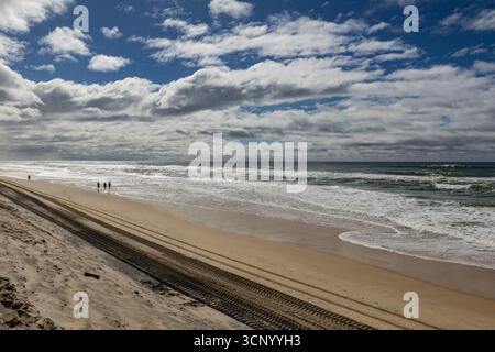 Ein breiter Sandstrand mit Meereswellen, die unter einem dramatischen Himmel an Land Rollen. Reifenspuren erstrecken sich entlang des Sandes, während ein paar Leute in der Nähe des Wassers spazieren Stockfoto