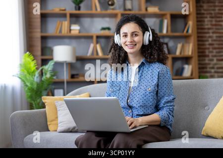 Eine junge Frau mit lockigem Haar, die weiße Kopfhörer und ein Jeansshirt trägt, lächelt in die Kamera, während sie auf einem Laptop schreibt, sitzt auf einem Sofa in einem modernen Wohnzimmer Stockfoto