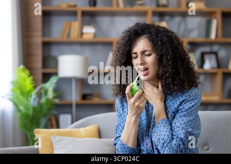 Junge Frau auf einer Couch, die Rachenmedizin in ihren Mund sprüht, zuckt vor Unwohlsein, während sie sich selbst behandelt Halsschmerzen und Erkältungssymptome zu Hause Stockfoto