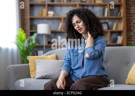 Junge Frau mit lockigem Haar, die sich unwohl fühlt und ihren schmerzhaften Nacken berührt, während sie zu Hause auf einem Sofa sitzt und Muskelverspannungen, Stress und Müdigkeit erlebt Stockfoto