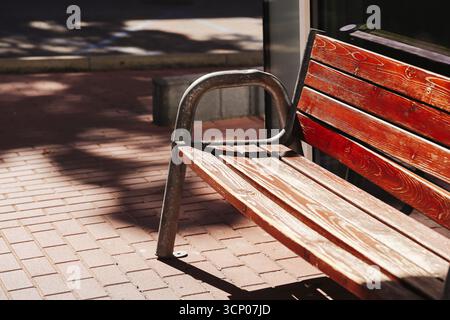 Holzbank mit eiserner Armlehne auf Ziegelpflaster. Straßenmöbel im Freien im Sonnenlicht. Ruheplatz im Park. Städtische Architektur. Stockfoto