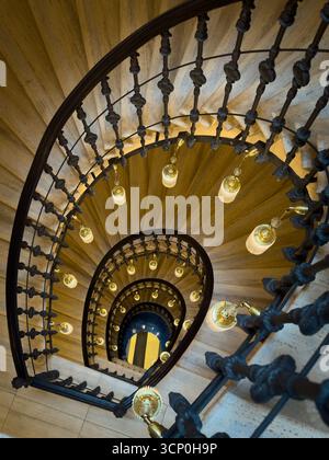 Wendeltreppe mit kunstvollem Eisengeländer und warmen Pendelleuchten von oben im historischen Innenraum Stockfoto