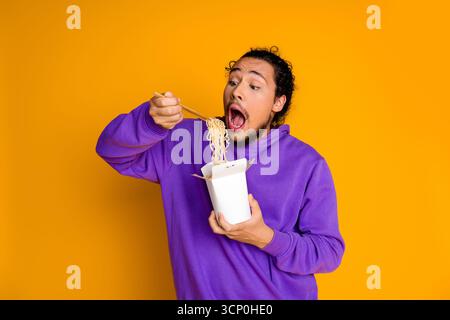 Junger Mann in violettem Hoodie, der Nudeln mit Essstäbchen auf einem leuchtend gelben Hintergrund genießt Stockfoto