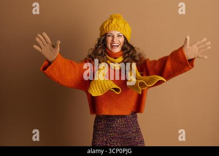 Eine junge Frau mit lockigen braunen Haaren, in einem warmen, orangen Pullover, gelbem Hut, Schal und Handschuhen, lächelt mit ausgestreckten Armen und begeistert Stockfoto