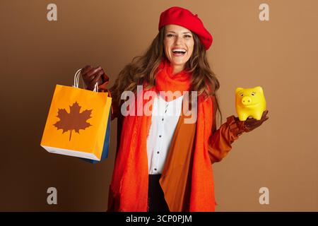 Eine fröhliche Frau in herbstlicher Mode, trägt Baskenmütze und orangefarbenen Mantel, lächelt, während sie Einkaufstaschen hält und gelbe Schweinebank. Das Bild erinnert an Themen Stockfoto