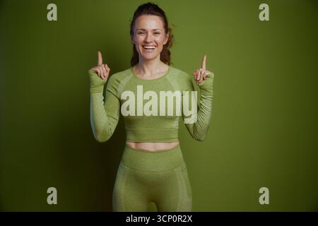 Eine lächelnde Frau in grüner Sportbekleidung zeigt nach oben, projiziert Fröhlichkeit und energetische Förderung von Fitness und Wohlbefinden gegen ein hellgrünes Stockfoto