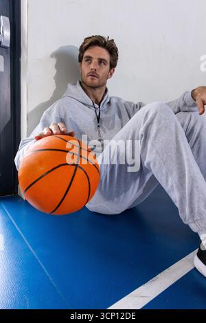 Mann in den späten Zwanzigern mit Trainingsanzug und Pfeife, der auf dem Court sitzt und Basketball hält Stockfoto