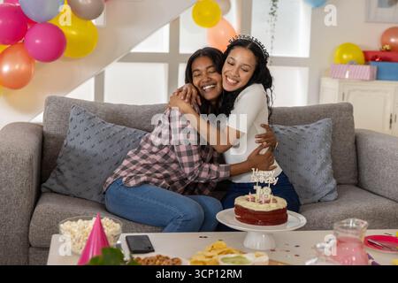 Diverse Mutter und Tochter im Teenageralter umarmen sich auf der grauen Couch im Wohnzimmer neben Geburtstagskuchen Stockfoto