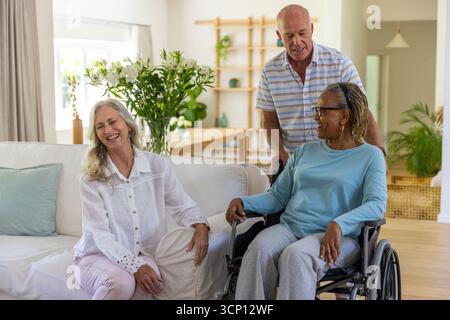 Verschiedene Seniorenfreunde lachen im Wohnzimmer mit Rollstuhl neben Sofa, Tisch, Kopierraum Stockfoto