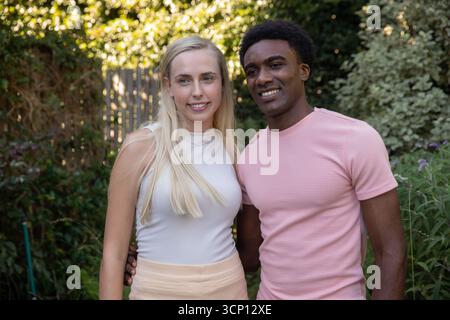 Weiße Frau und afroamerikanisches Männerpaar stehen im sonnigen Garten und tragen ein cremefarbenes T-Shirt Stockfoto