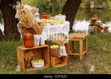 Eine rustikale Herbstszene mit einem dekorierten Tisch im Freien an einem ruhigen Gewässer. Der Tisch, mit einem weißen Tuch bedeckt, ist mit einer schönen Decke beladen Stockfoto