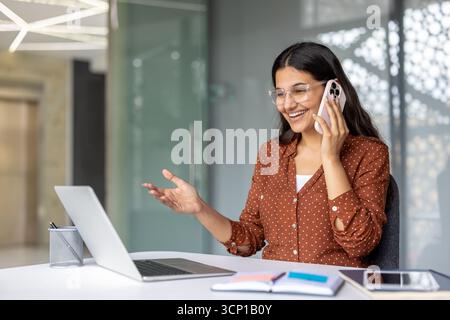 Junge indianerin in Brillen, die sich am Mobiltelefon unterhalten und mit der Hand Gesten machen kann, während sie in moderner Büroumgebung am Laptop arbeitet Stockfoto