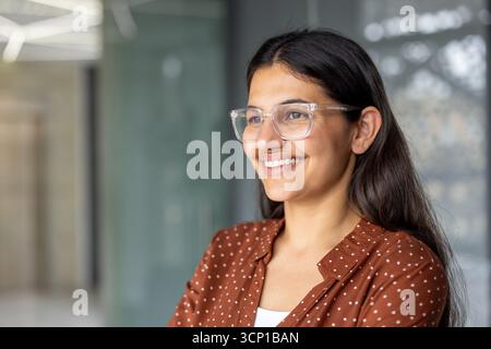 Junge indische Profi-Frau in Klarsichtbrille und brauner Polka Dot-Bluse, die selbstbewusst lächelt, während sie in einer modernen Büroumgebung steht, Kopfaufnahme von der Seite Stockfoto