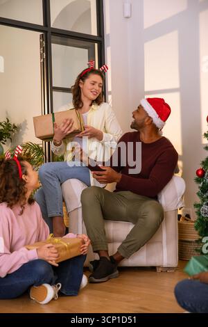 Verschiedene Familien tauschen Geschenkverpackungen aus, tragen weihnachtsmütze, Stirnbänder am weihnachtsbaum zu Hause Stockfoto