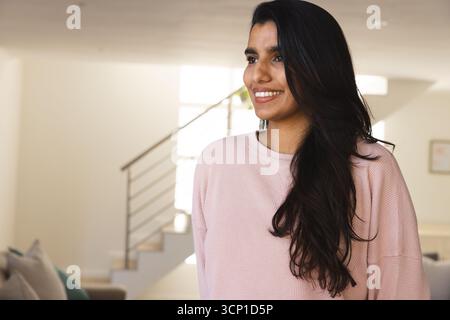 Asiatische Frau lächelt und steht im modernen Wohnzimmer mit rosa Pullover in der Nähe der Treppe Stockfoto