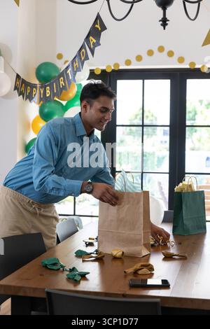 Asiatische Männer arrangieren Geschenktüten auf Esstisch zu Hause unter Happy Birthday Banner, Ballons Stockfoto