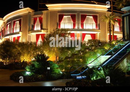 Luxuriöses beleuchtetes Gebäude in Las Vegas bei Nacht mit eleganten roten Vorhängen, üppigem Grün, Palmen und einer leuchtenden Rolltreppe im Freien Stockfoto
