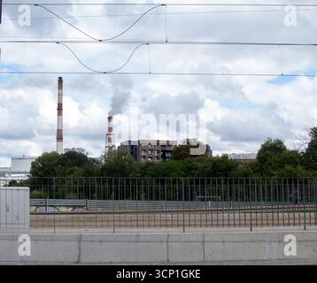 Rauch steigt aus Industrieschornsteinen in einem städtischen Gebiet mit bewölktem Himmel und Grün in der Nähe Stockfoto
