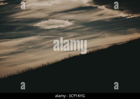 Dramatischer Himmel über einem Silhouettenhang. Ein beeindruckendes Bild zeigt eine Silhouette am Hang vor einem dynamischen Himmel voller strukturierter Wolken und schafft so Stockfoto