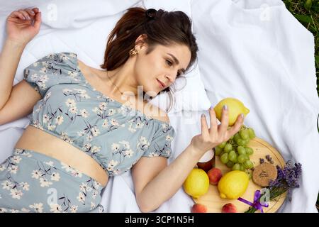 Junge Frau, die auf einer Picknickdecke im Sommerkleid liegt und Zitrone hält, umgeben von Früchten, Trauben, Pfirsichen, Lavendel, Outdoor-Lifestyle und Entspannung Stockfoto