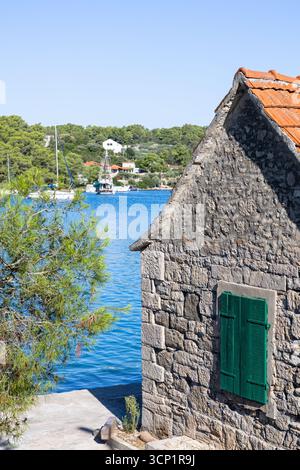Ein rustikales Steinhaus mit hellen grünen Fensterläden liegt neben einer ruhigen Küstenbucht. Boote ruhen im blauen Hafen, während entfernte Häuser und Kiefern Stockfoto