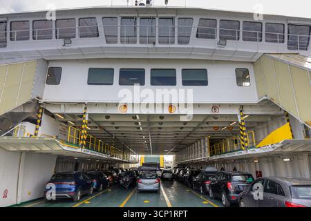 Ein belebtes Autodeck auf einer großen Passagierfähre, gefüllt mit geparkten Autos unter dem Oberdeck. Geländer und gelbe Sicherheitsmarkierungen säumen die Seiten, konv Stockfoto
