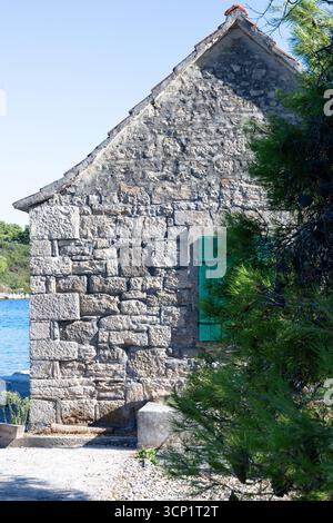 Ein rustikales Steinhaus befindet sich am Ufer des Wassers mit leuchtenden grünen Fensterläden. Ruhiges blaues Wasser, Kiefern und weit entfernte Boote schaffen eine friedliche Küste Stockfoto