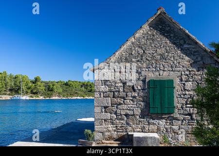 Ein rustikales Steinhaus befindet sich am Ufer des Wassers mit leuchtenden grünen Fensterläden. Ruhiges blaues Wasser, Kiefern und weit entfernte Boote schaffen eine friedliche Küste Stockfoto