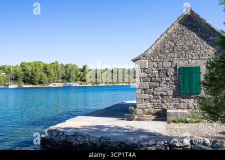 Ein rustikales Steinhaus befindet sich am Ufer des Wassers mit leuchtenden grünen Fensterläden. Ruhiges blaues Wasser, Kiefern und weit entfernte Boote schaffen eine friedliche Küste Stockfoto