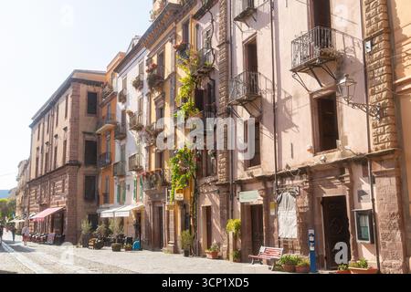 Historische Straße in Sardinien mit farbenfrohen, rustikalen Fassaden von Gebäuden mit Balkonen und Blumen. Ein paar Leute schlendern gemütlich entlang Stockfoto