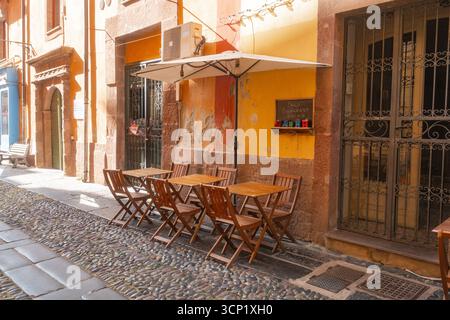 Leere Holztische und -Stühle sind vor einem malerischen Café in einer malerischen Kopfsteinpflasterstraße angeordnet, die Besucher einladen, sich in einer einladenden Atmosphäre zu entspannen Stockfoto