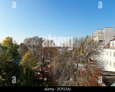 Wien, Österreich - 22. Oktober 2024: Herbstbäume säumen einen Innenhof neben modernen weißen Gebäuden und Sitzgelegenheiten im Freien unter hellblauem Himmel in Wi Stockfoto