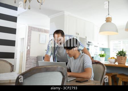 Asiatischer Vater, der den Sohn am Küchentisch mit Laptop und Smartphone führt Stockfoto