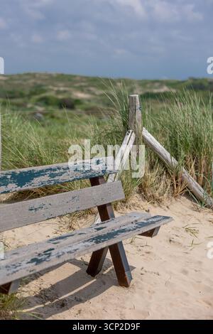 Verwitterte Holzbank auf einer Sanddüne, mit Blick auf eine natürliche grasbewachsene Landschaft unter einem bewölkten Himmel, die an Konzepte von Einsamkeit, Entspannung, a erinnert Stockfoto