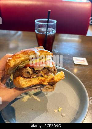 Halb gegessen Burger, garniert mit Salat, Tomate und Zwiebeln, gehalten von einer Hand über einem grauen Teller in einem Restaurant mit einem verschwommenen Getränk. Stockfoto