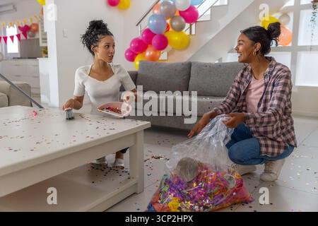 Vielfältige Mutter und Teenager-Tochter, die Konfetti in rosa Staubpfanne und Tasche im Wohnzimmer fegen Stockfoto