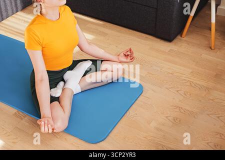 Unerkennbare Frau in einem gelben Hemd und grünen Shorts sitzt in Lotusposition auf einer blauen Yogamatte. Stockfoto