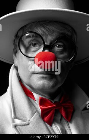 Nahaufnahme selektives Farbporträt des traurigen Clowns. Ein älterer Mann in einem weißen Mantel und Hut auf schwarzem Hintergrund. Stockfoto