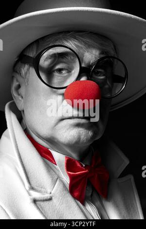 Nahaufnahme selektives Farbporträt des traurigen Clowns. Ein älterer Mann in einem weißen Mantel und Hut auf schwarzem Hintergrund. Stockfoto