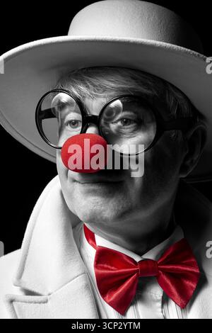 Nahaufnahme selektives Farbporträt des traurigen Clowns. Ein älterer Mann in einem weißen Mantel und Hut auf schwarzem Hintergrund. Stockfoto