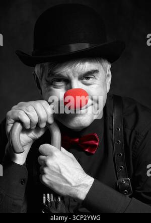 Selektives Farbporträt eines lachenden Clowns am Griff eines Schirms. Ein älterer Mann mit roter Clownnnase und Bowler-Hut auf dem Kopf. ich war angezogen Stockfoto